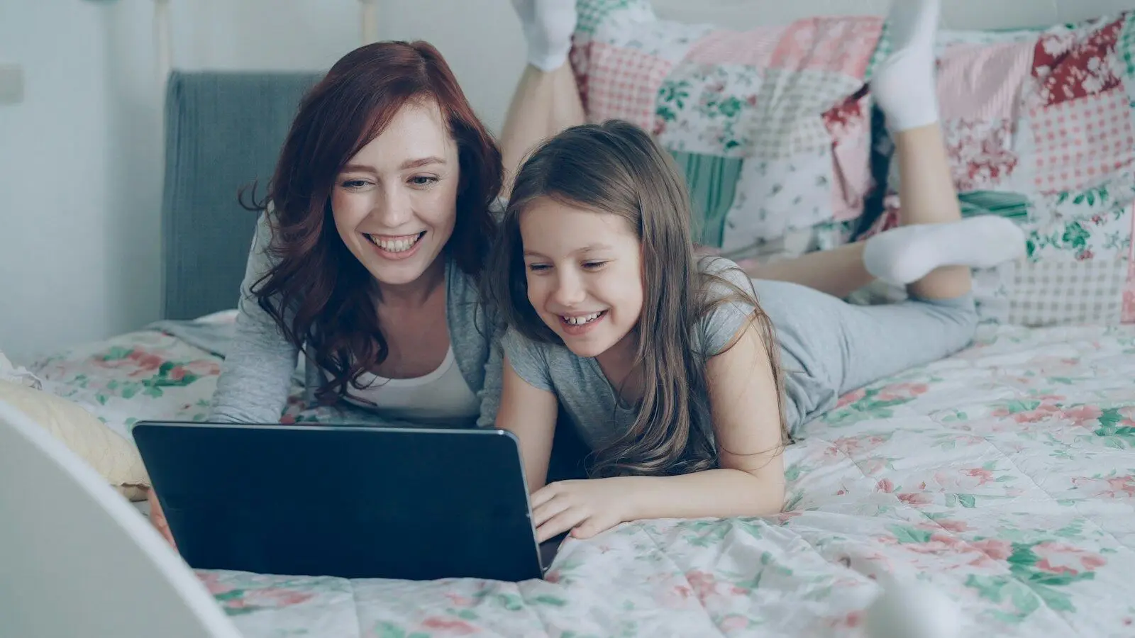 Mãe e filha deitadas na cama, sorrindo enquanto olham juntas para um notebook